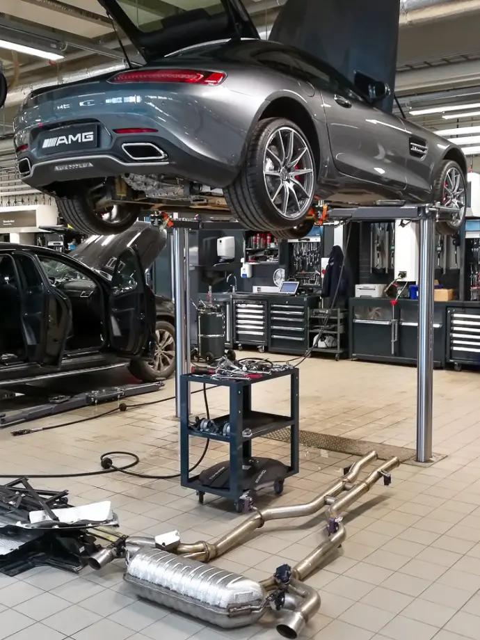 Mercedes-Benz merkkihuolto Kuvassa AMG mersu nosturilla ylös nostettuna ja konepelti auki. Maassa kiiltävä tupla-pakoputkisto, ja auton alla työkalukärry täynnä osia ja työkaluja.