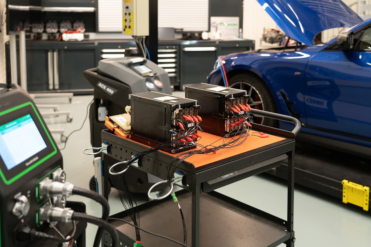 Siireltävän vaunun päällä kaksi moduulia sähköauton akustosta. Moduulien kennoihin kytketty useita mittausjohtumia. Taustalla sininen BMW ilmastointihuollossa.
