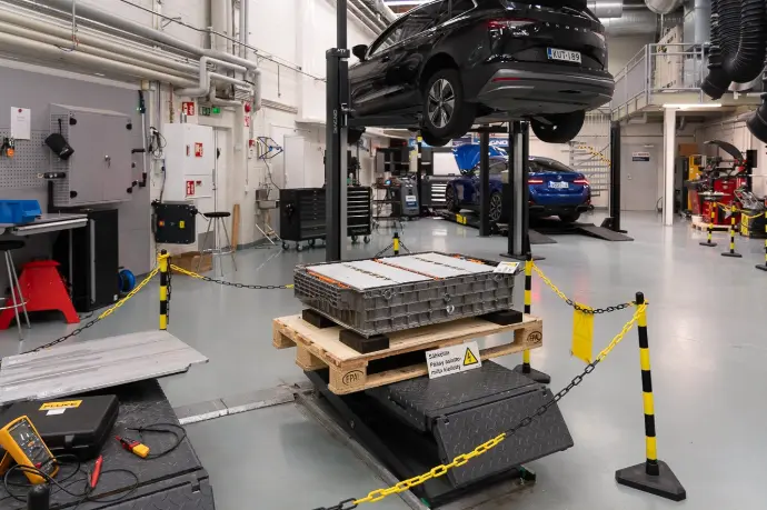 Kuvassa kolmas nostin n. metrin korkeudelle nostettuna, jonka päällä korkeajänniteakku ja jännitetyökaluja. Alue rajattu musta-keltaisilla tolpilla ja ketjulla. Taustalla Skoda 2-pilarinostimella ylös nostettuna ja BMW 4-pilarinostimella ilmastointihuollossa.