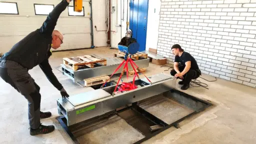 Kaksi Diagnon huoltoasentajaa asentamassa lattiaan upotettavaa jarrudynamometri-iskunvaimennintesteri kompleksia. Moduulia asetetaan paikalleen nostolaitteen avulla ja se kohdistetaan käsin.